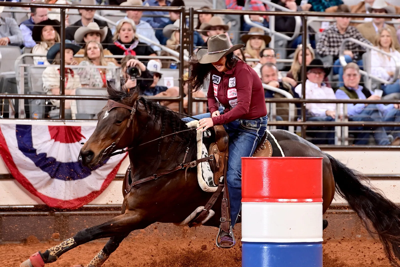 A barrel racer and horse speeding around a barrel in an arena.